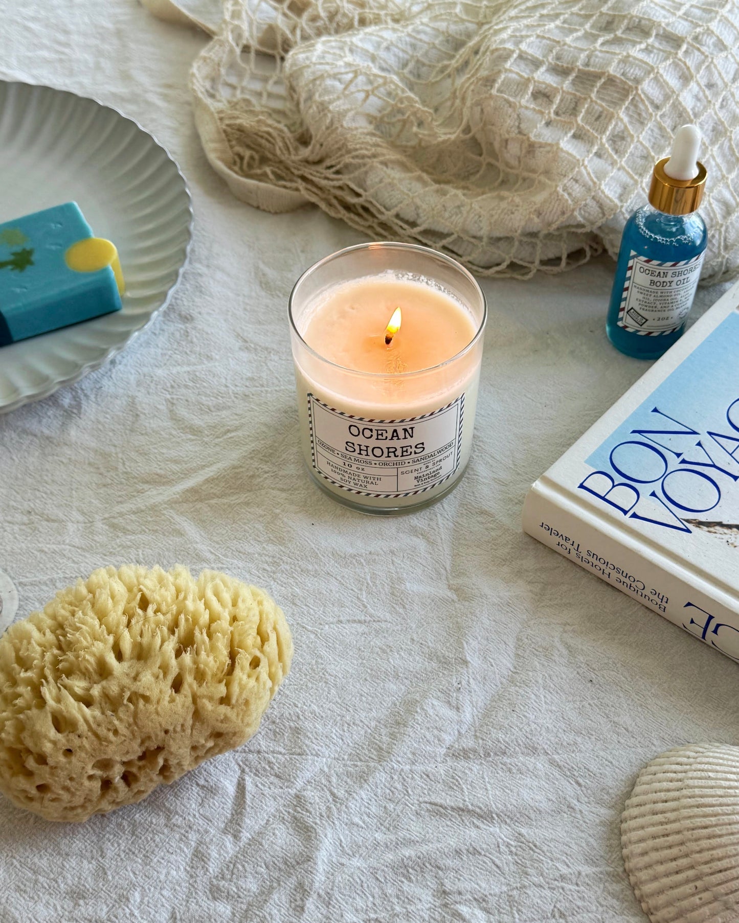 Candle labeled 'Ocean Shores' with a sponge, shell, and book on a textured surface