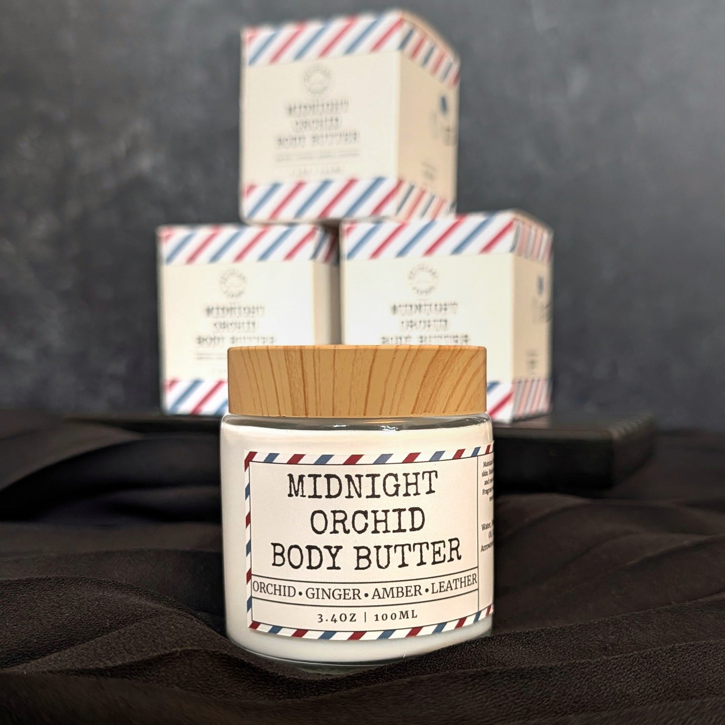 Jar of Midnight Orchid Body Butter with packaging in the background on a dark surface.