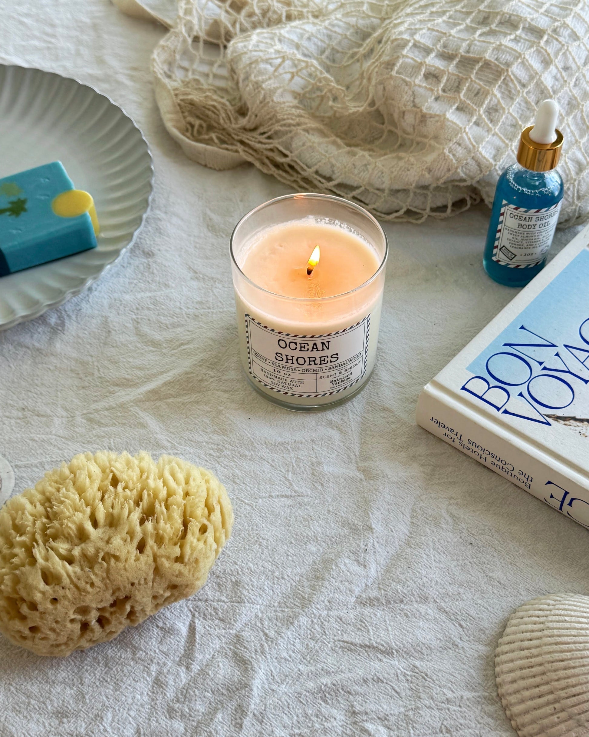 Candle labeled 'Ocean Shores' with a sponge, shell, and book on a textured surface
