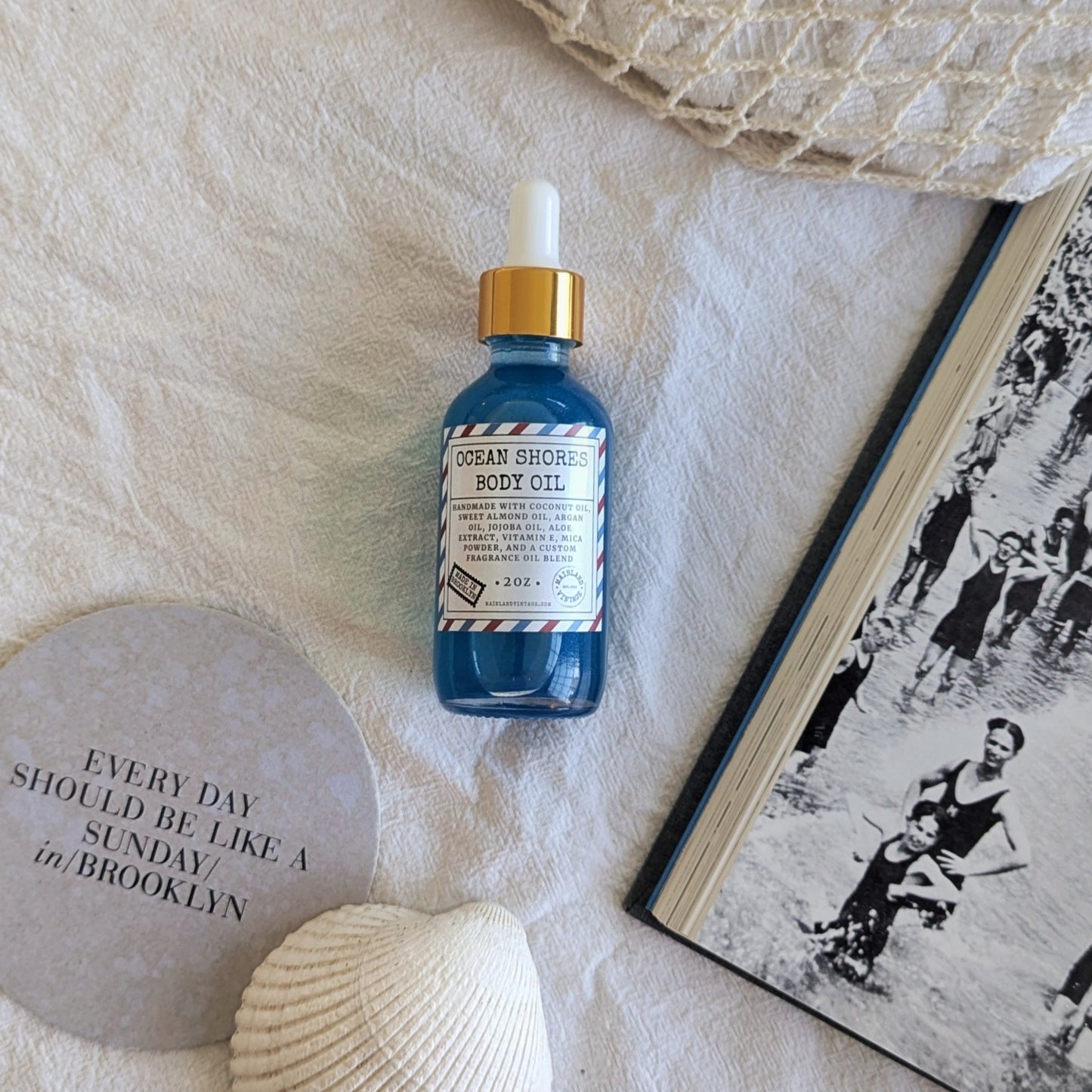 Blue bottle of Ocean Shores Body Oil on a textured surface with a shell and book.