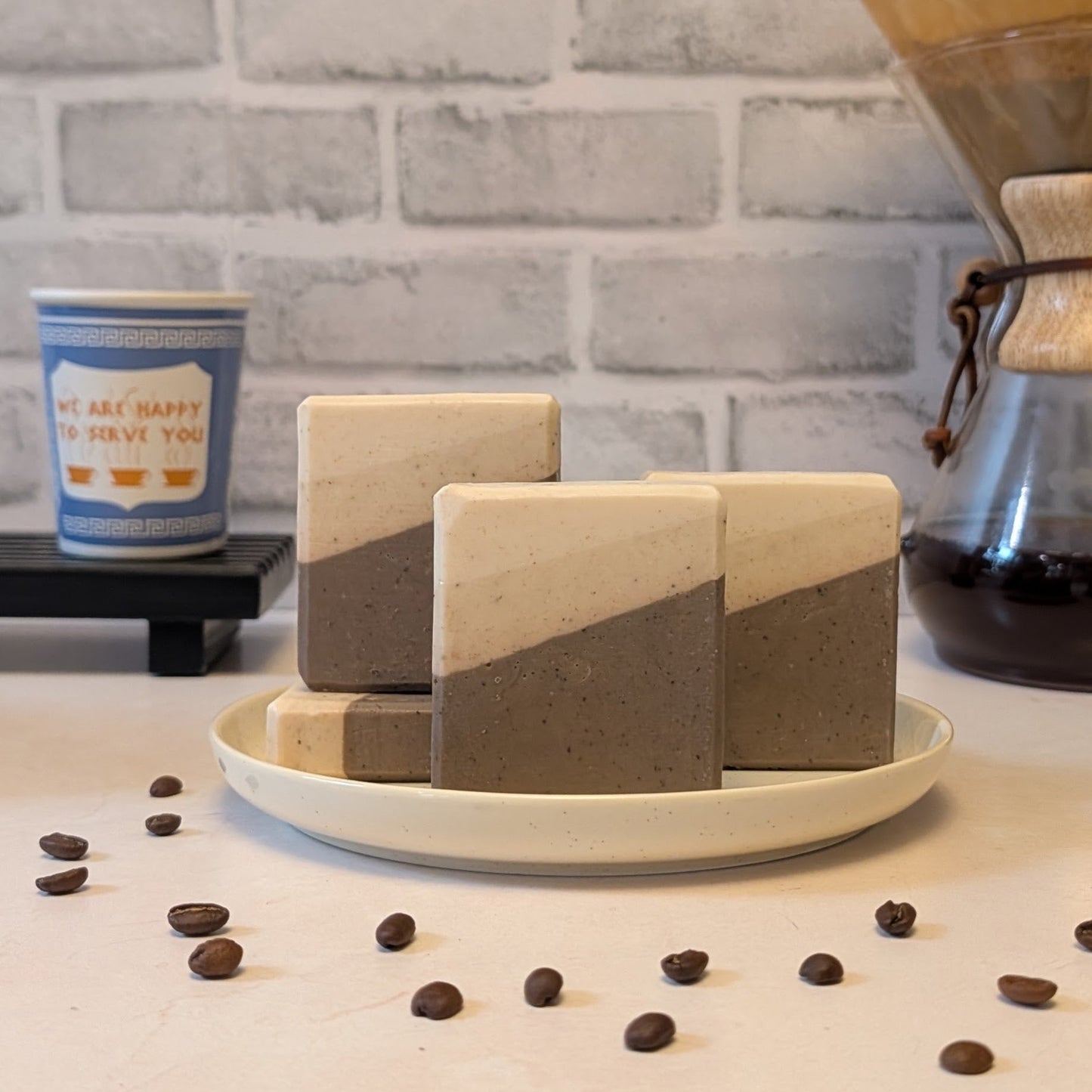 Three geometrically shaped soap bars on a white plate with coffee beans and a coffee cup in the background.
