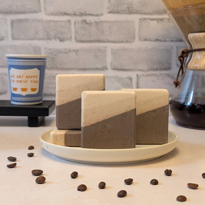 Three geometrically shaped soap bars on a white plate with coffee beans and a coffee cup in the background.