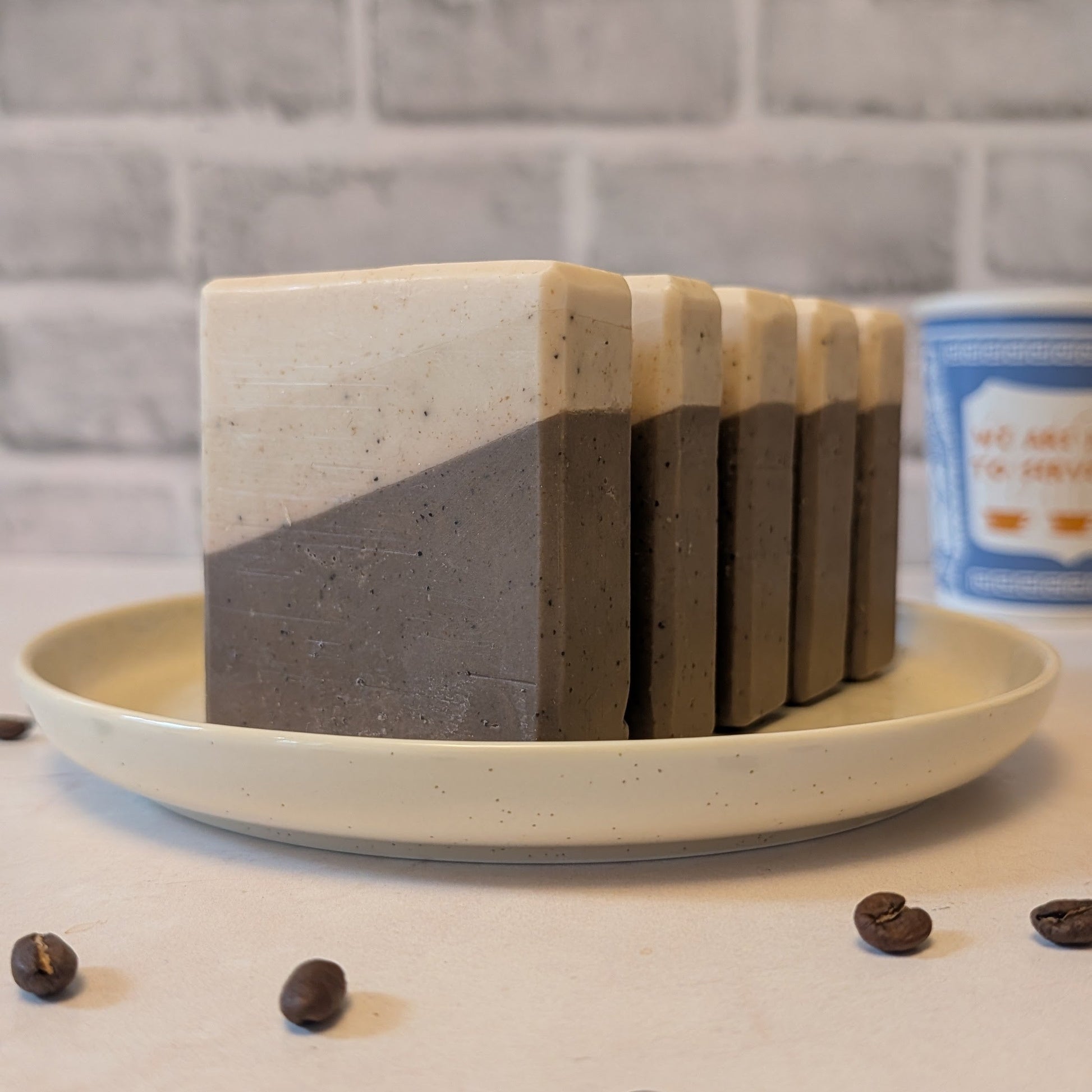 Stack of layered soap bars on a plate with coffee beans around, against a brick wall background.