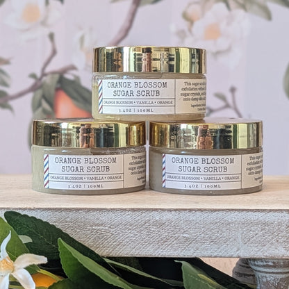 Three jars of Orange Blossom Sugar Scrub stacked on a wooden surface with a floral background.