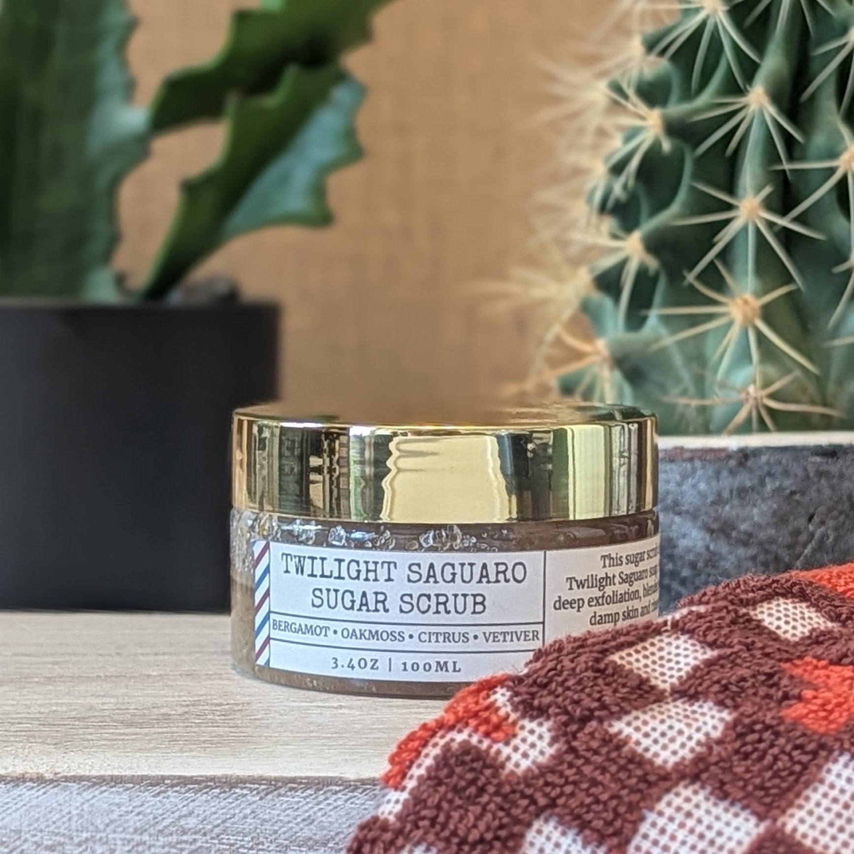 Twilight Saguaro Sugar Scrub jar with gold lid on a surface with plants in the background