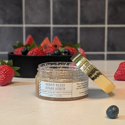 A jar of Berry Bliss Sugar Scrub with its gold lid leaning beside it on a light countertop. Fresh strawberries, blueberries, and watermelon slices are arranged around the jar, with a dark tiled backsplash behind it.