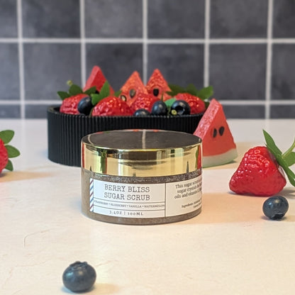 A jar of Berry Bliss Sugar Scrub sits on a light countertop with strawberries, blueberries, and watermelon slices arranged around it, and a tiled wall in the background.