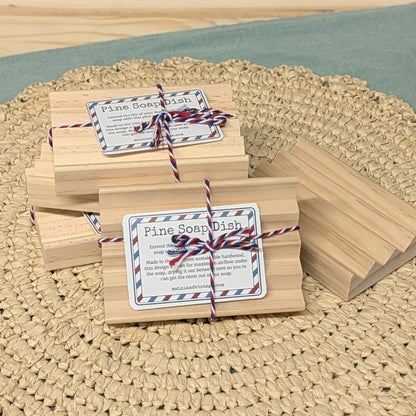 Two small wooden boxes tied with red-and-white string and a white label reading 'Pine Soap Dish' are arranged on a round woven beige placemat over a muted green surface.