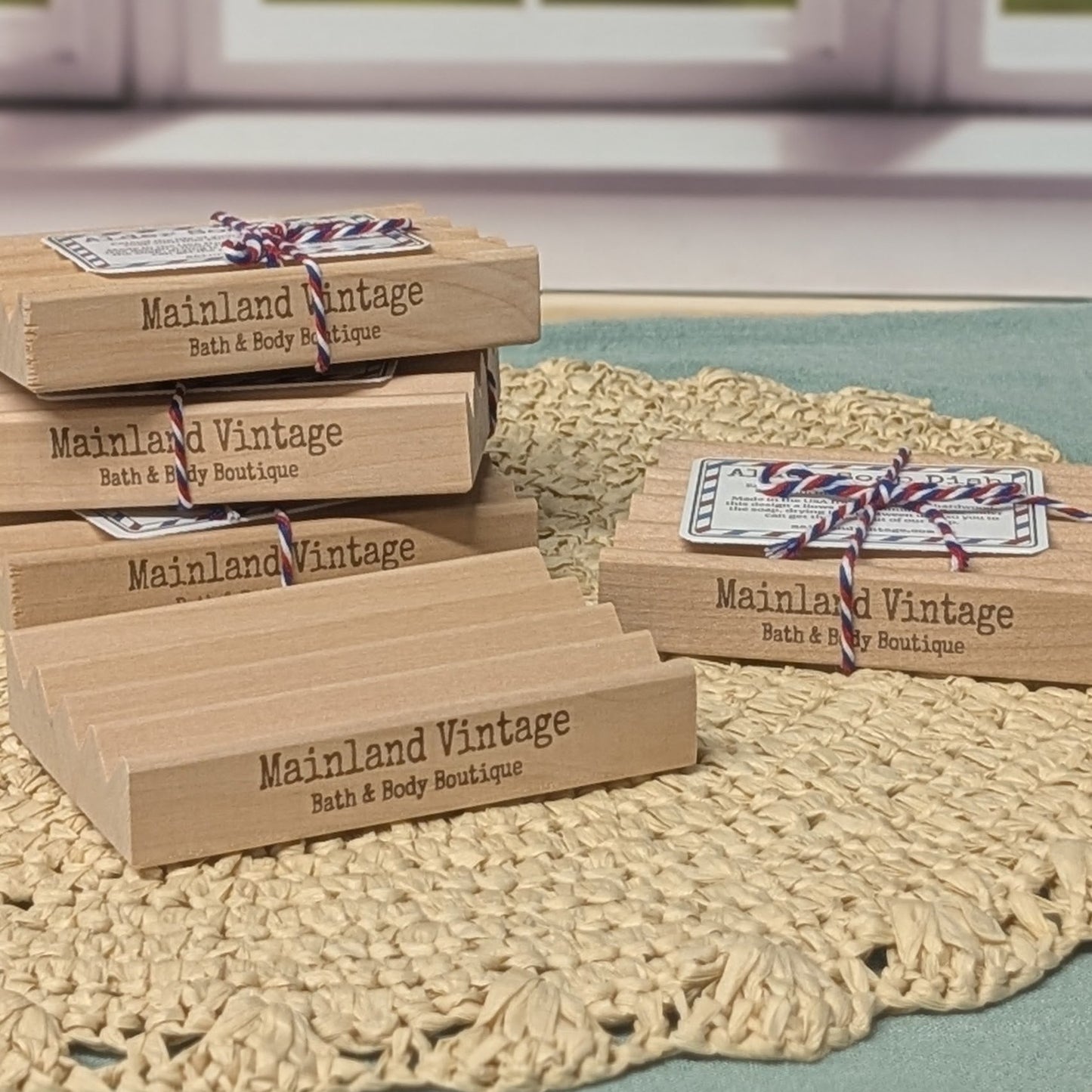 Several rectangular wooden gift boxes engraved with the brand name 'Mainland Vintage Bath & Body Boutique' are arranged on a woven beige doily over a green surface, with a softly blurred window in the background.