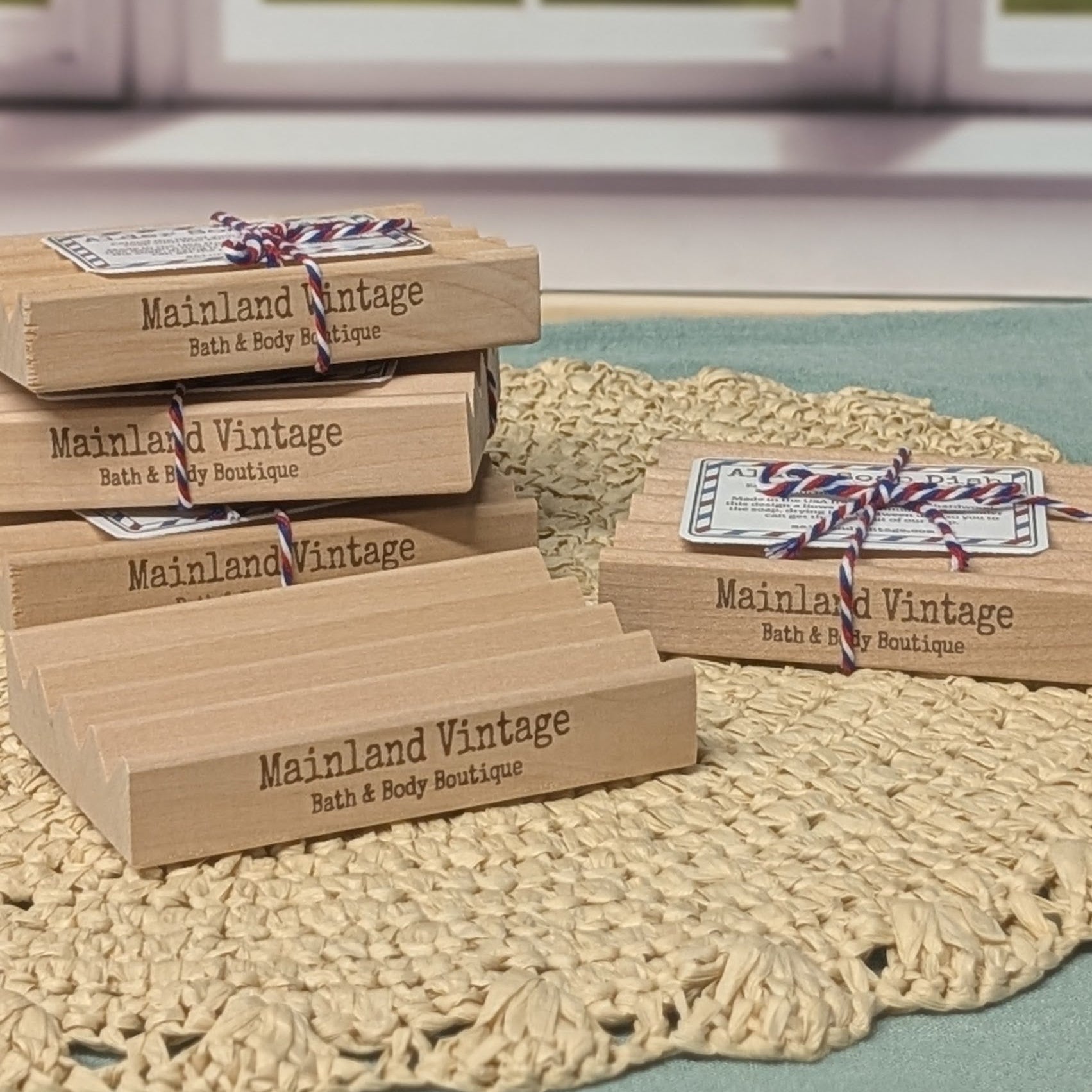 Several rectangular wooden gift boxes engraved with the brand name 'Mainland Vintage Bath & Body Boutique' are arranged on a woven beige doily over a green surface, with a softly blurred window in the background.