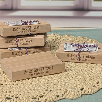 Several rectangular wooden gift boxes engraved with the brand name 'Mainland Vintage Bath & Body Boutique' are arranged on a woven beige doily over a green surface, with a softly blurred window in the background.