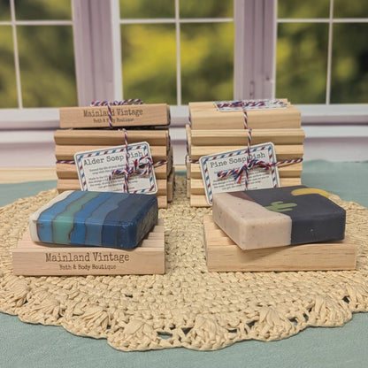 Several bars of handmade bath and body soap displayed on small wooden blocks over a crocheted doily, with a window and blurred greenery in the background. Visible labels include Mainland Vintage, Elderberry Bath & Body, Pine Scent Bath & Body, and Elderberry.