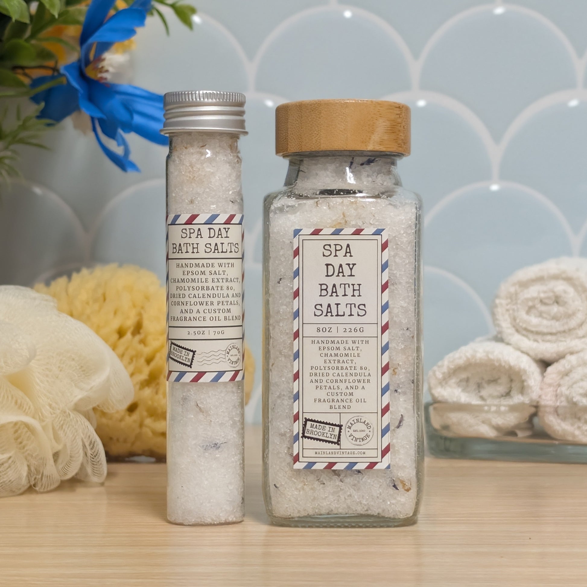 Two containers of spa day bath salts on a light wood surface against a blue scalloped tile wall. A clear glass jar with a bamboo lid and a clear glass tube with a silver cap are both labeled 'SPA DAY BATH SALTS'.