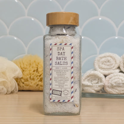 A clear glass bottle of spa day bath salts with a wooden lid and a vintage-style label, set on a light wood surface with a blue scalloped tile background and bath accessories.