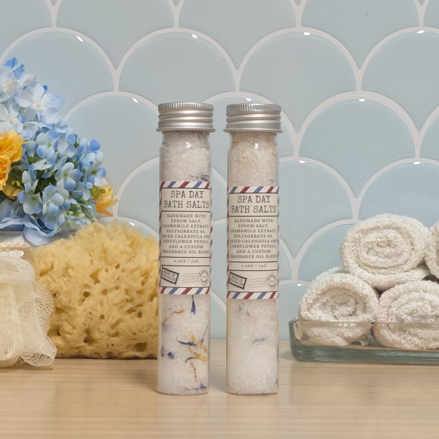 Two glass vials of bath salts labeled 'SPA DAY BATH SALTS'  with silver caps, displayed on a wooden surface with a blue scalloped tile background, a sponge, flowers, and rolled towels.