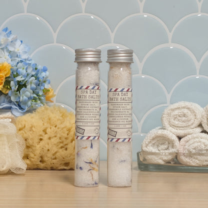 Two glass vials of bath salts labeled 'SPA DAY BATH SALTS'  with silver caps, displayed on a wooden surface with a blue scalloped tile background, a sponge, flowers, and rolled towels.