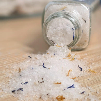 White crystal bath salts with dried blue and yellow flowers spilling from a clear glass bottle onto a light wooden surface.