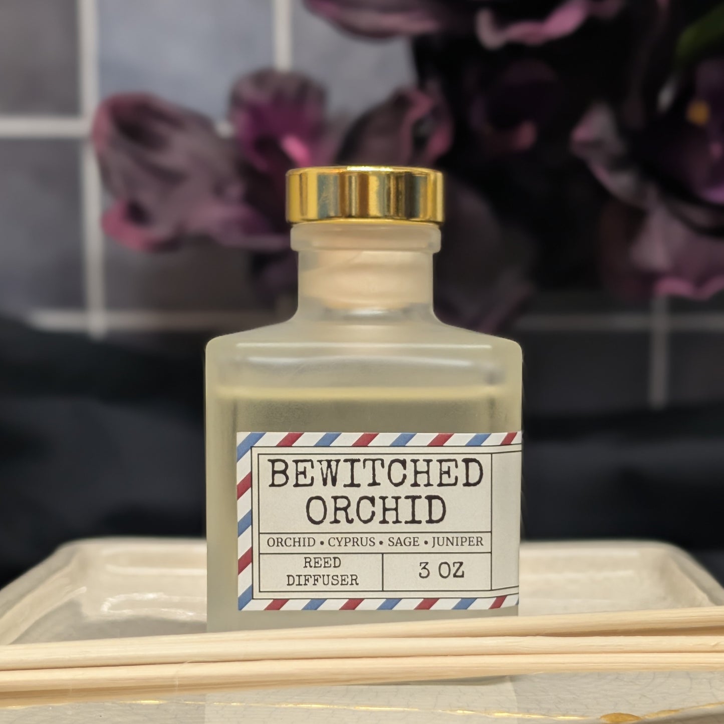 A small frosted glass diffuser bottle with a gold cap and a label reading 'Bewitched Orchid,' set on a tray with reed sticks in the foreground and dark floral decor blurred in the background.