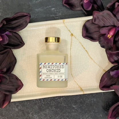 A frosted glass reed diffuser bottle labeled 'Bewitched Orchid' with a gold cap, placed on a white ceramic tray with gold veining, surrounded by dark purple orchid flowers on a dark gray textured surface.
