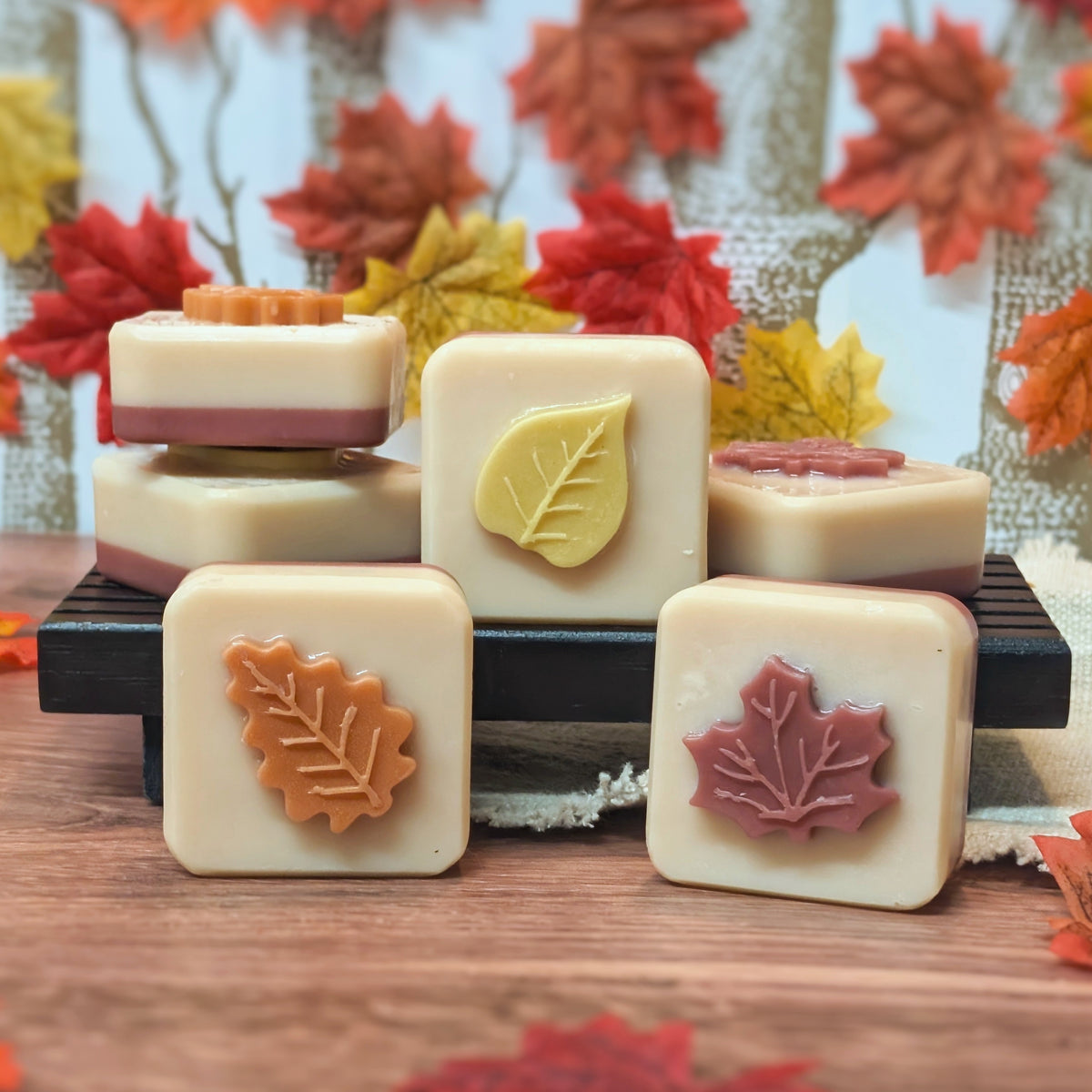 Six square cream-colored soap bars decorated with autumn-themed leaf designs, arranged on a black tray with a rustic background of colorful artificial leaves.