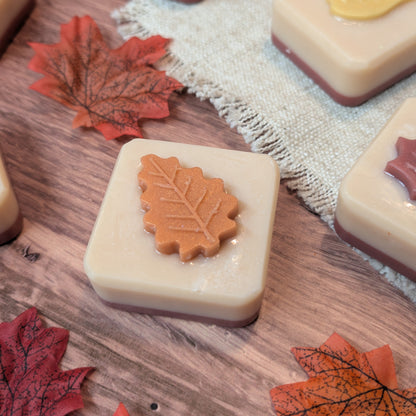 Square beige soap bars with embossed autumn leaf designs on top, arranged on a wooden surface with faux maple leaves and a burlap cloth.