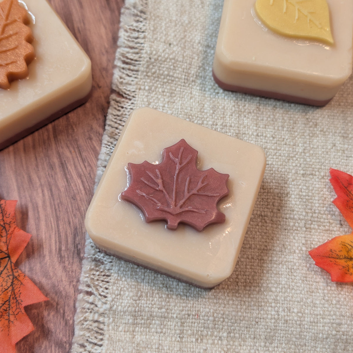 Three square beige soap bars with embossed autumn leaf designs are arranged on a textured linen cloth with scattered faux maple leaves. One soap has a brown leaf, one has a reddish-brown leaf, and one has a yellow leaf.