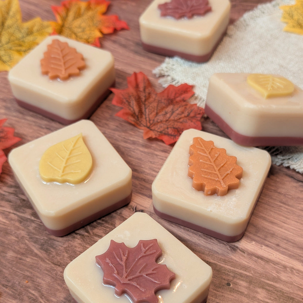 Six square cream-colored soap bars decorated with autumn leaf motifs in orange, red, and yellow, arranged on a rustic wooden surface with scattered decorative leaves.