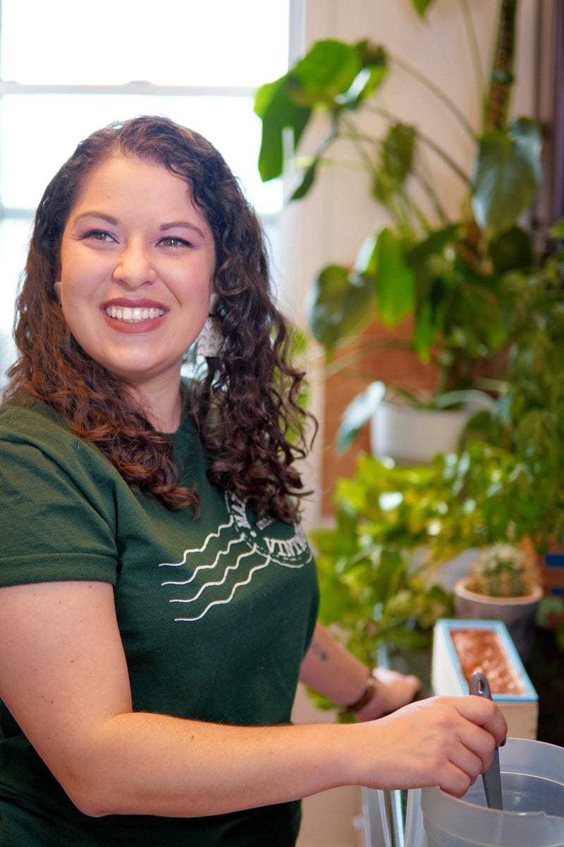 Woman standing looking to the side. Wearing a green shirt with the Mainland Vintage logo. On the counter in front of her is soap she is making. Behind her are plants